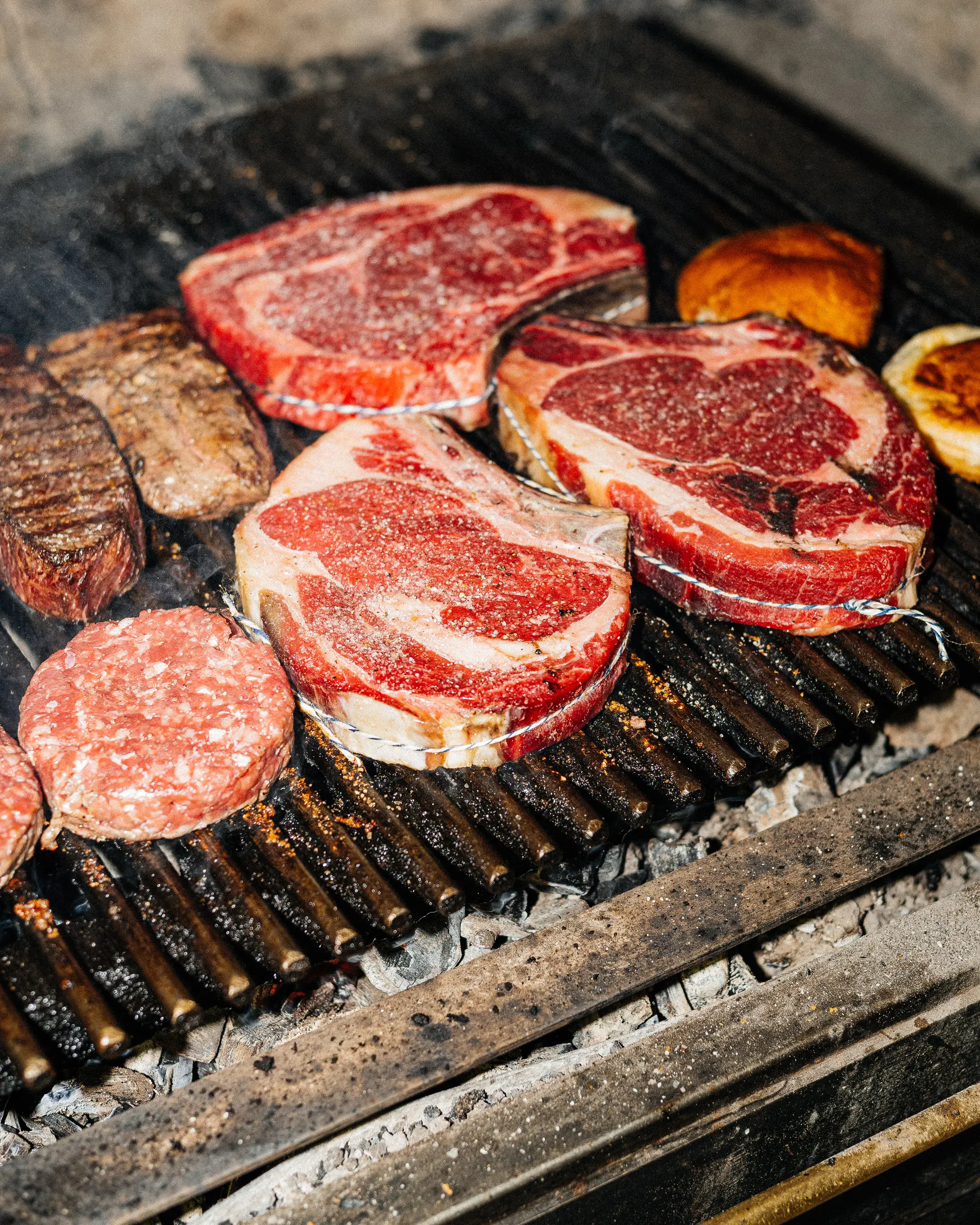 Hovězí steaky a burger na grilu připravované na otevřeném ohni.