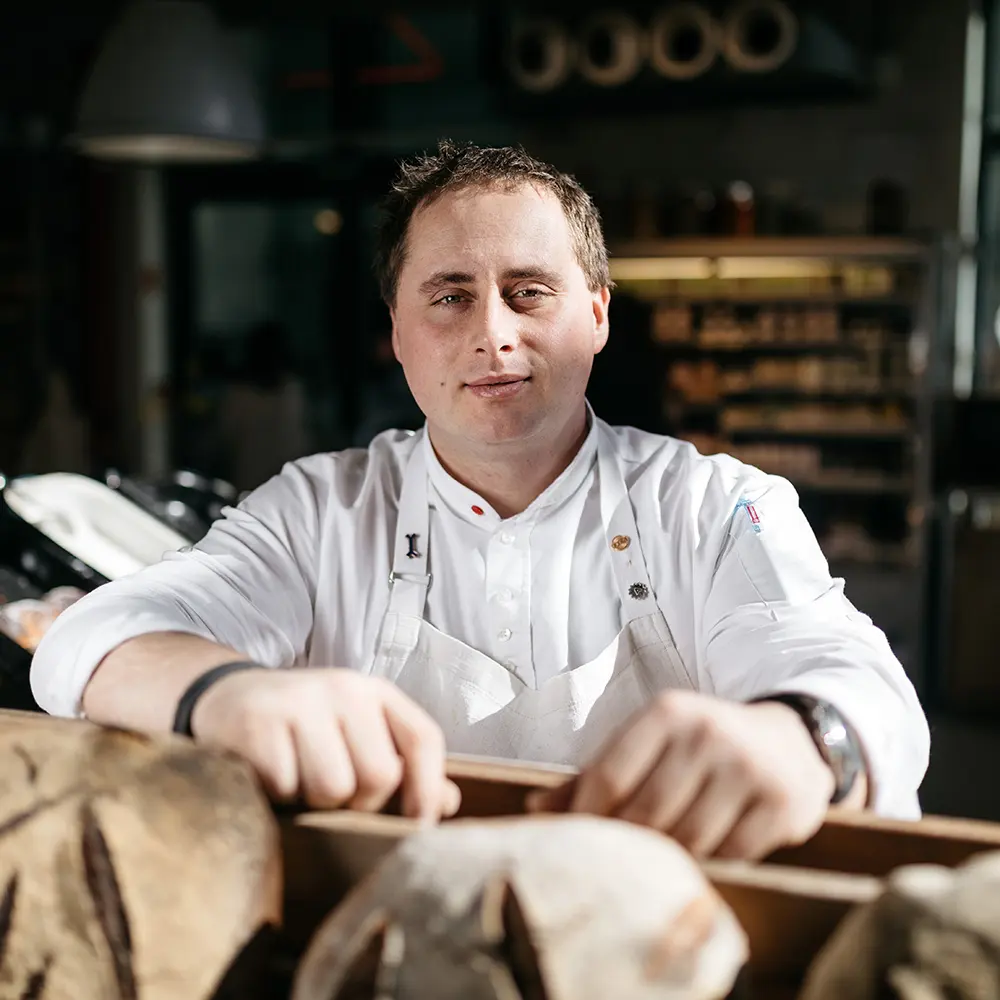 Baker in a white apron with fresh bread loaves in a bakery.