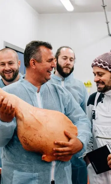 Group of men in protective gowns examining a large pork leg and laughing.