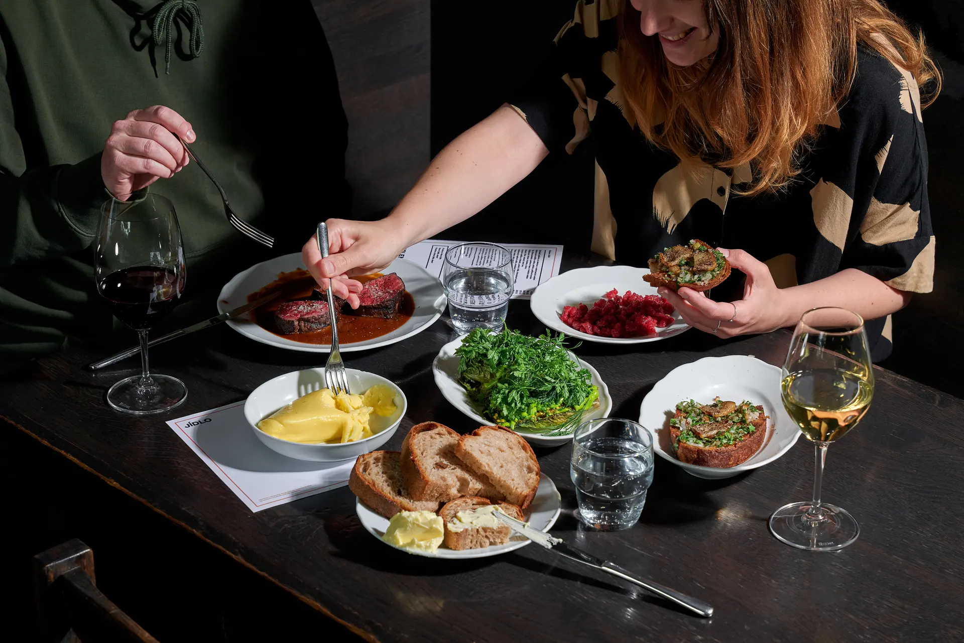 Dvojice hostů sdílí večeři se steakem, přílohami a vínem u tmavého stolu v restauraci.