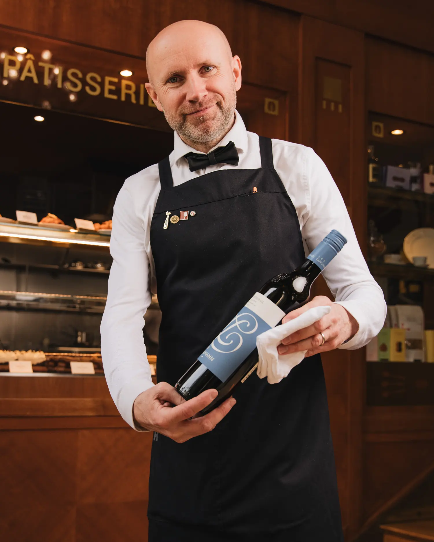 Sommelier holding a bottle of red wine in front of a dessert display in a restaurant.