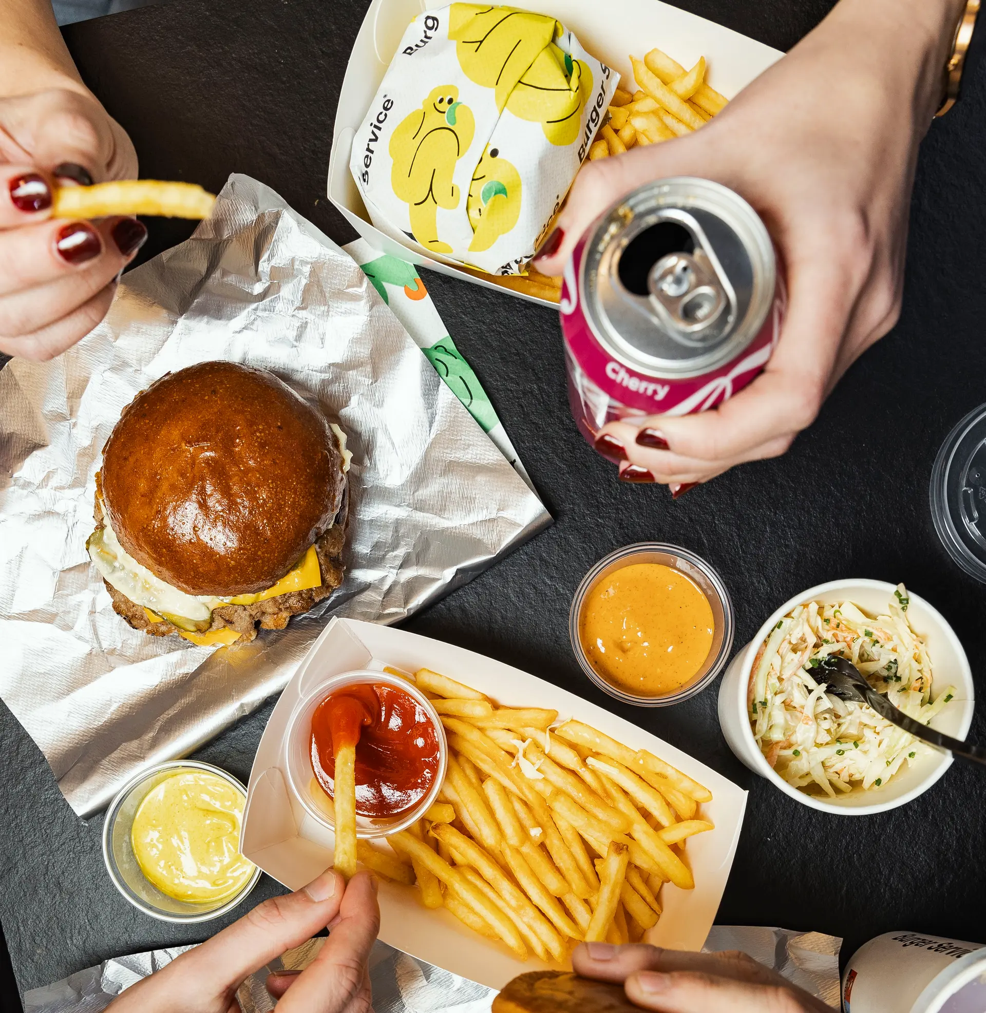 Burger s hranolkami, dipy a limonádou na stole, ruce lidí při společném jídle.