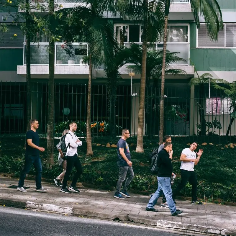 Skupina lidí kráčející po městském chodníku v rezidenční čtvrti se zelení a palmami.